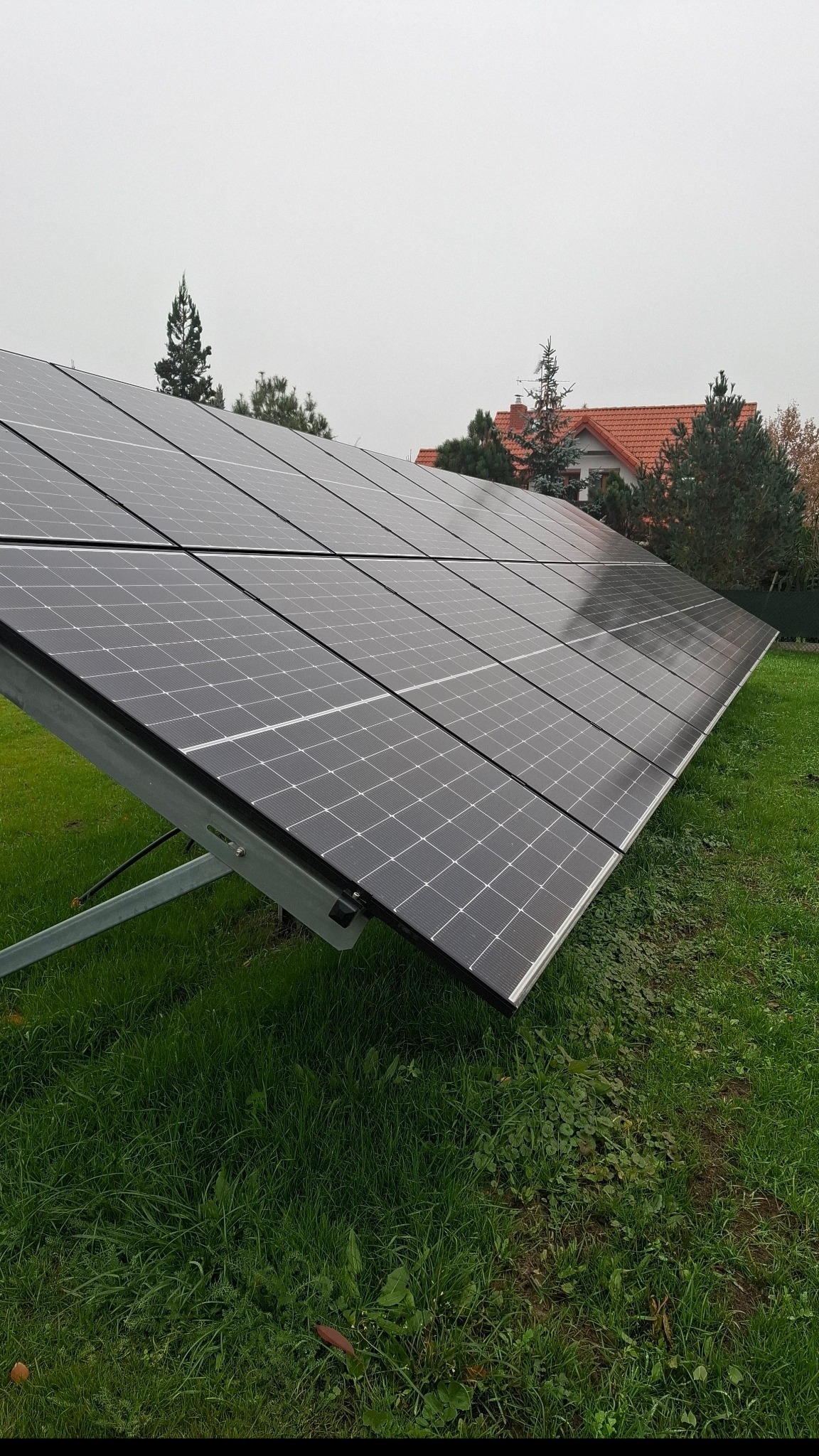 Rząd paneli słonecznych na metalowej konstrukcji, zainstalowany na trawniku przed domem z czerwoną dachówką w pochmurny dzień.