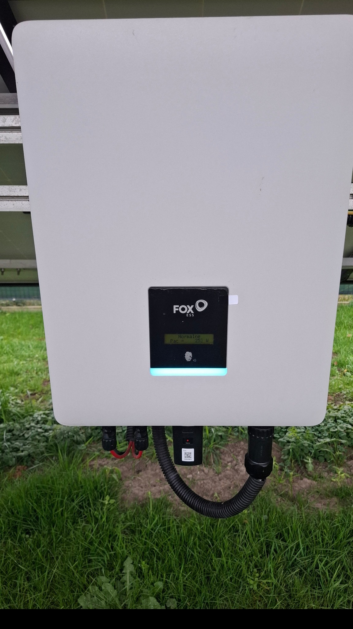 Biała obudowa inwertera Fox ESS z wyświetlaczem wskazującym moc 252W, zamontowana na tle zielonej trawy, widoczne kable i złącza.