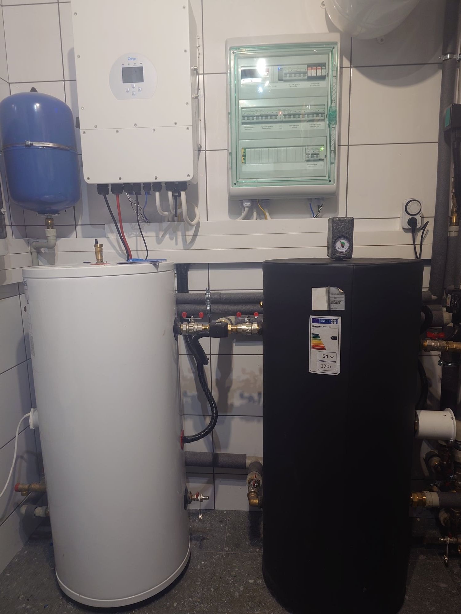 Wnętrze kotłowni z białym zbiornikiem akumulacyjnym, czarnym bojlerem, białym inwerterem i rozdzielnicą elektryczną na ścianie z białych płytek.