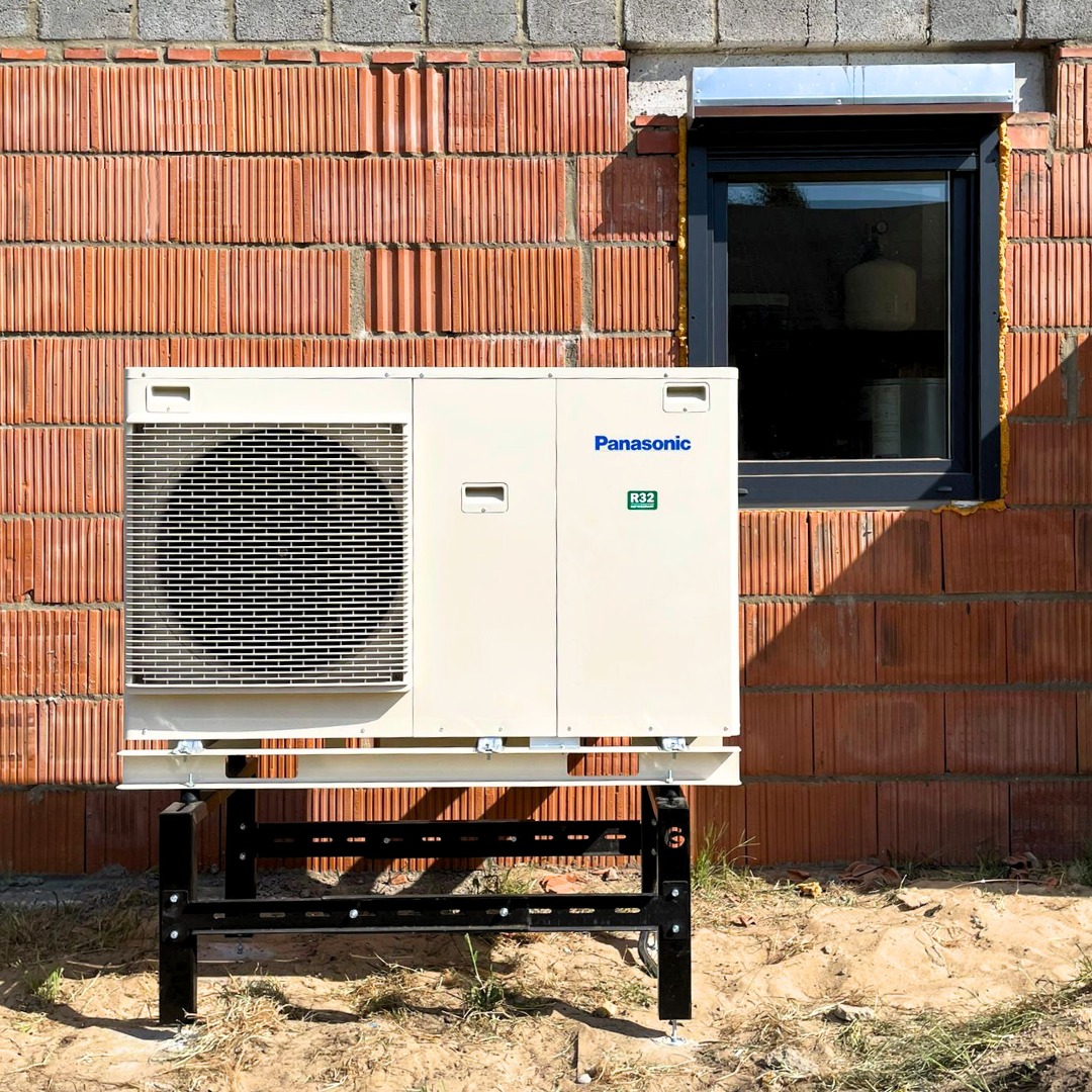 Zewnętrzna jednostka pompy ciepła Panasonic Aquarea zamontowana na czarnej, metalowej konstrukcji przed ścianą z czerwonej cegły w surowym stanie, obok okna z szarą ramą.