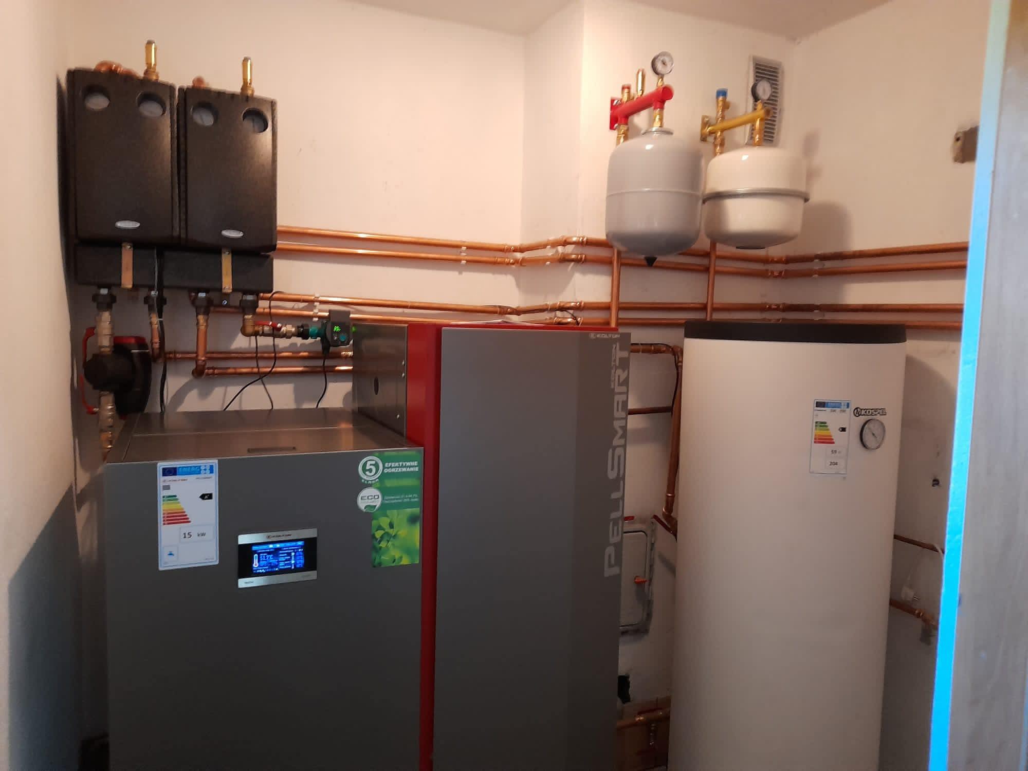 Kompletna instalacja kotła na pellet z zasobnikiem ciepłej wody użytkowej, widoczne rury miedziane, zbiorniki wyrównawcze i sterownik. Kocioł marki PellSmart.