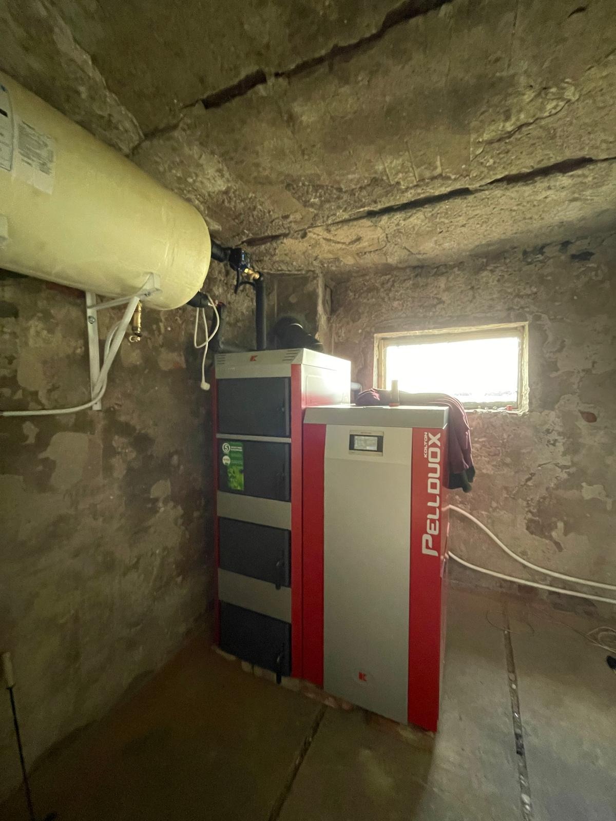 Montaż kotła na pellet Kolton Pellduox w piwnicy w Łodzi, widoczny zasobnik na wodę, surowe ściany i małe okno.