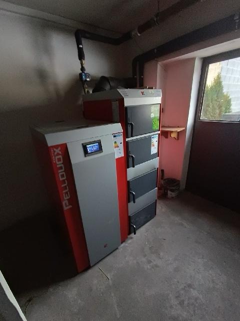 Kocioł na pellet Pellduox z widocznym wyświetlaczem i podajnikiem, zainstalowany w kotłowni z betonową podłogą i rurami grzewczymi pod sufitem.