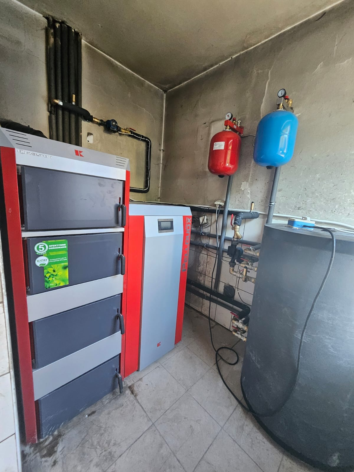 Kocioł na paliwo stałe z podajnikiem, widoczne zawory, rury i zbiorniki wyrównawcze w kolorze czerwonym i niebieskim, zainstalowane w kotłowni o surowych ścianach.