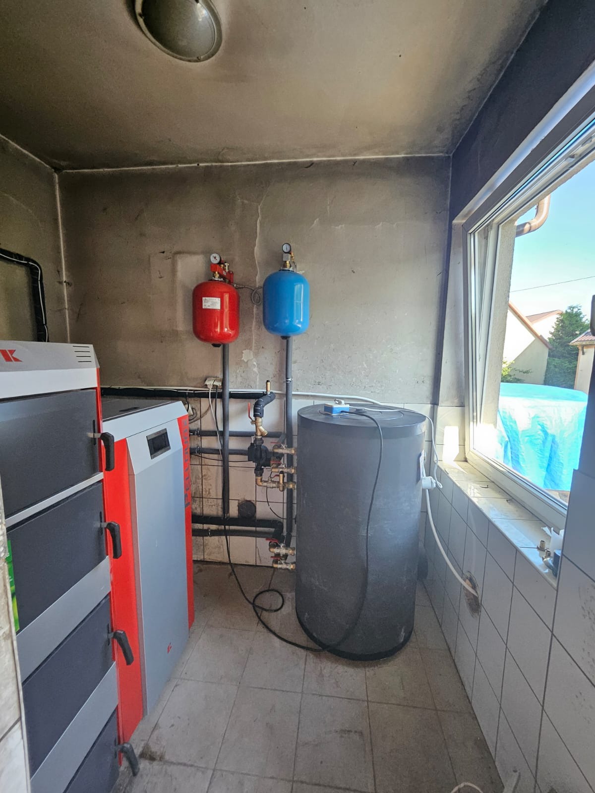 Kotłownia z piecem na paliwo stałe, zbiornikiem akumulacyjnym, czerwonym i niebieskim naczyniem przeponowym, widoczne instalacje hydrauliczne.