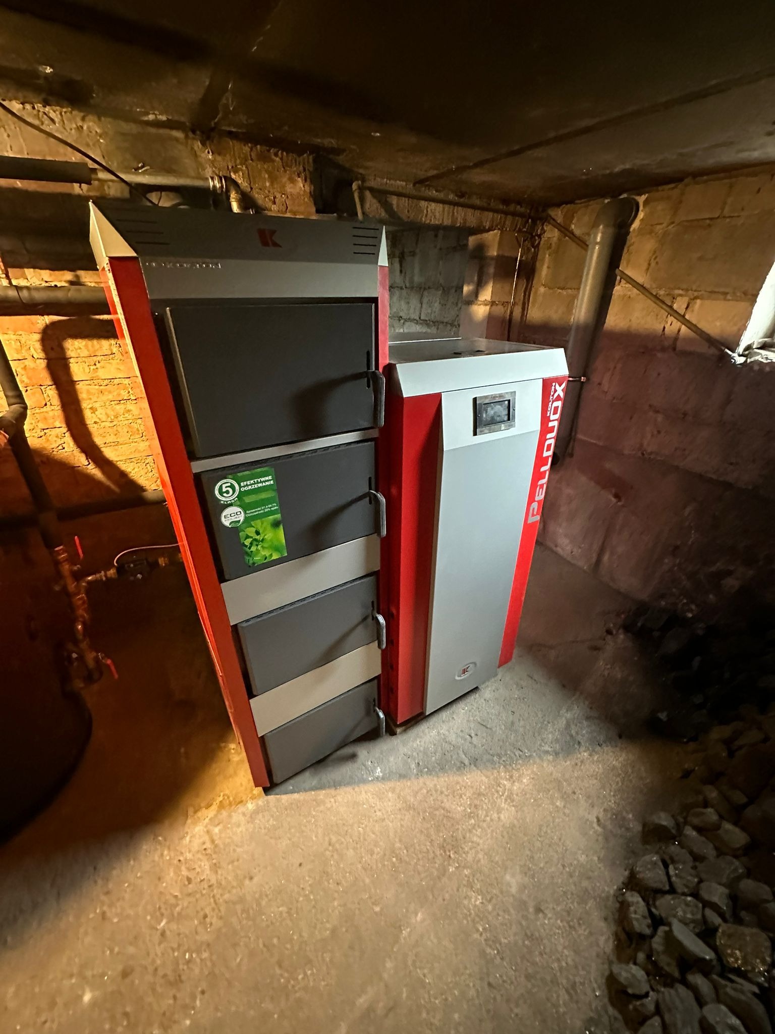 Kocioł na pellet z podajnikiem w piwnicy, widoczny certyfikat '5 klasa' i oznaczenie ECO, obok leżą brykiety węgla.