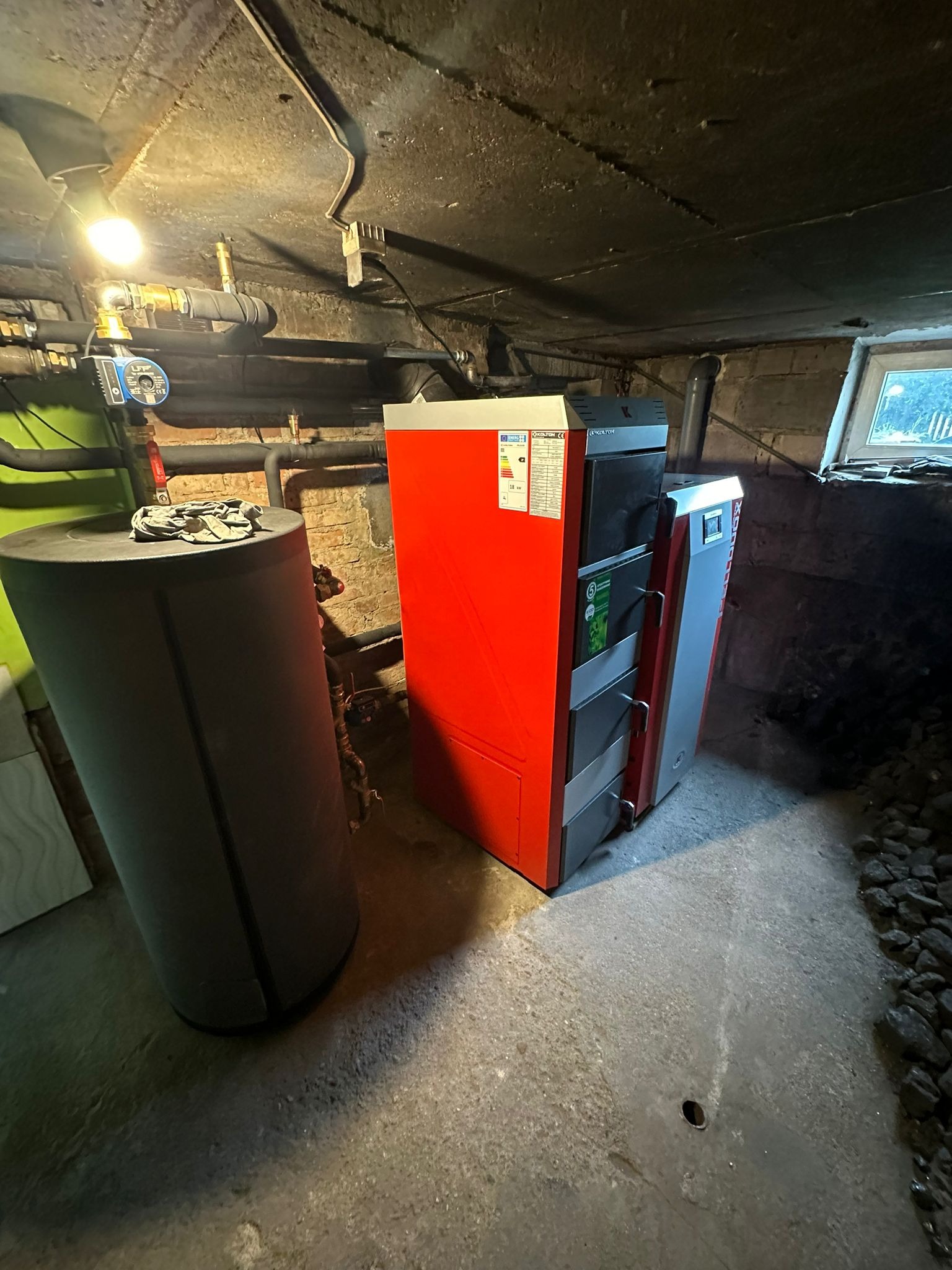 Kotłownia z kotłem na paliwo stałe Kolton w kolorze czerwonym i szarym, obok szary zbiornik akumulacyjny, widoczne instalacje grzewcze pod sufitem, w tle okno piwniczne.