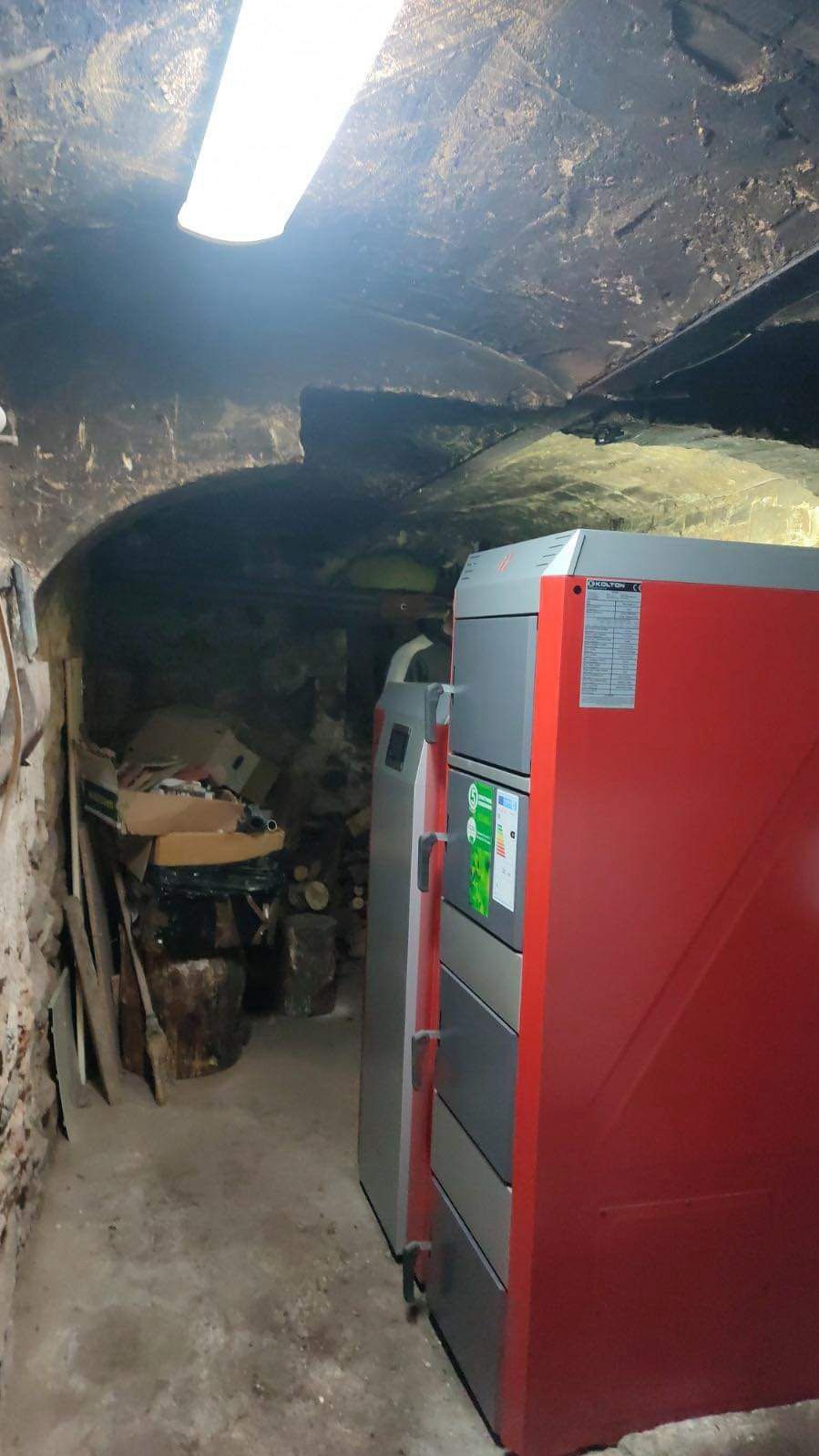 Czerwono-szary piec w piwnicy z łukowym sklepieniem, oświetlony jarzeniówką, obok składowane drewno i kartony.