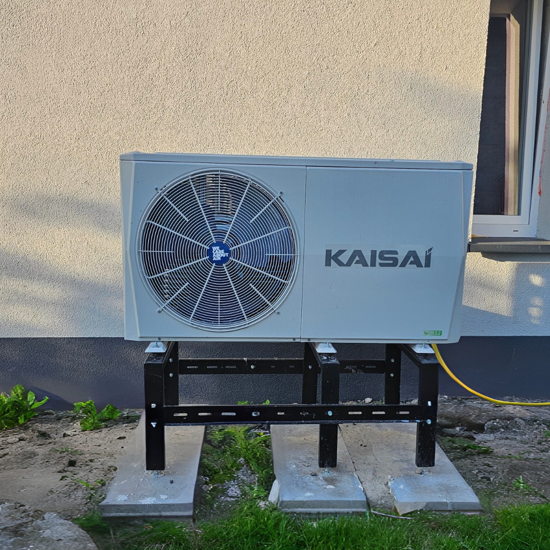Zewnętrzna jednostka pompy ciepła marki Kaisai, zamontowana na metalowej konstrukcji na betonowych płytach przed budynkiem z oknem.