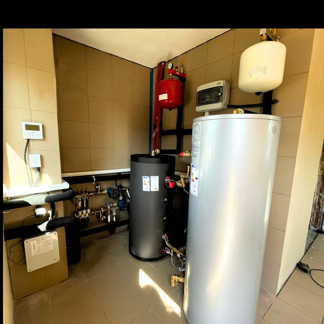 Kompaktowa kotłownia z dwoma zbiornikami akumulacyjnymi: szarym i srebrnym, połączonymi rurami, z czerwonym zbiornikiem wyrównawczym i sterownikiem na ścianie wyłożonej kafelkami.