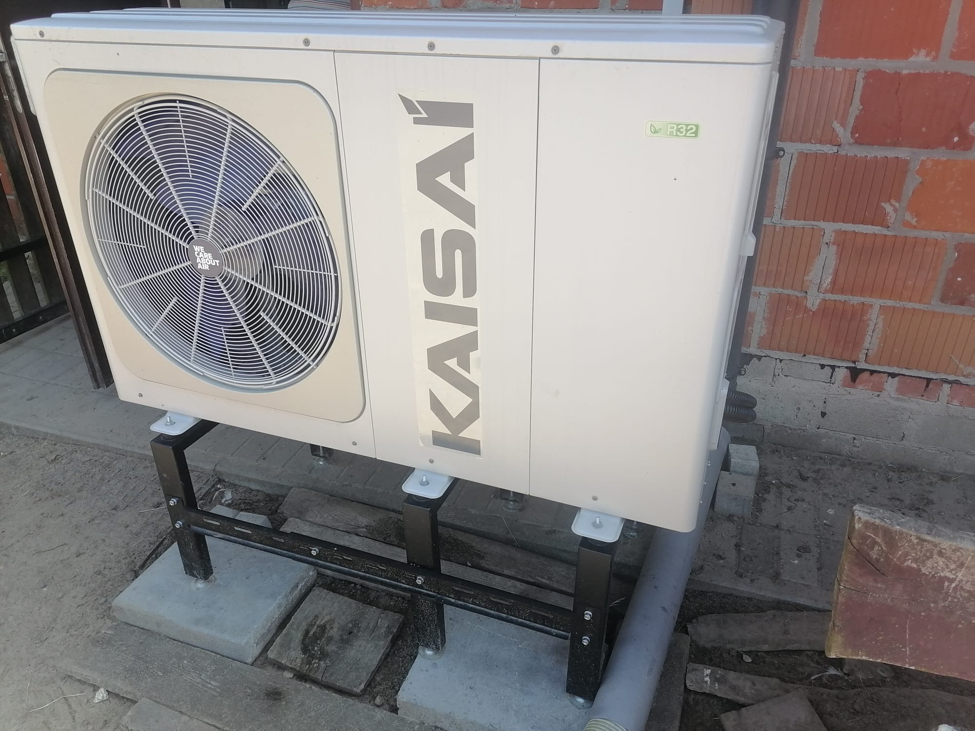 Zewnętrzna jednostka pompy ciepła marki Kaisai zamontowana na czarnej metalowej konstrukcji na betonowych płytach, z widoczną ścianą z czerwonej cegły w tle.