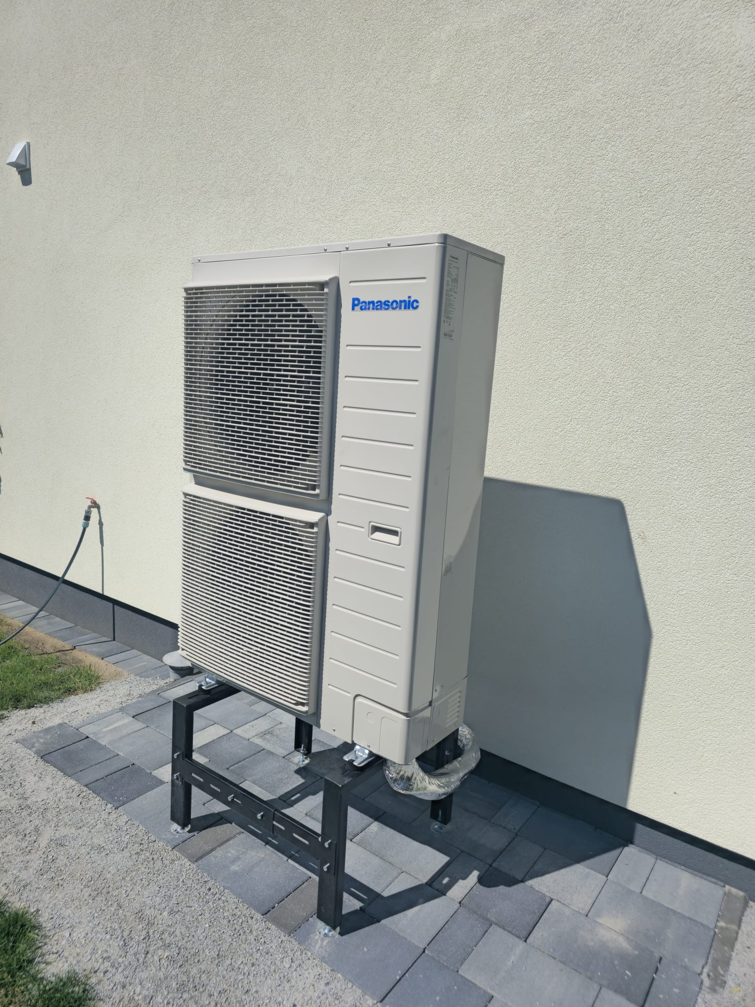 Zewnętrzna jednostka klimatyzacji Panasonic na czarnym, metalowym stelażu, ustawiona na szarej kostce brukowej przy jasnej ścianie budynku. Widoczne podłączenia rur w srebrnej izolacji.
