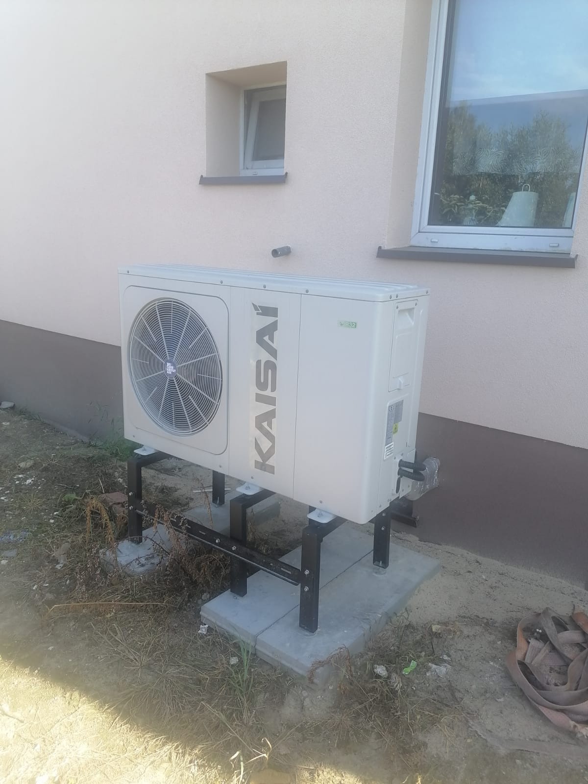 Zewnętrzna jednostka pompy ciepła Kaisai zamontowana na czarnej, metalowej konstrukcji na betonowych płytach przy jasnej ścianie budynku z dwoma oknami.