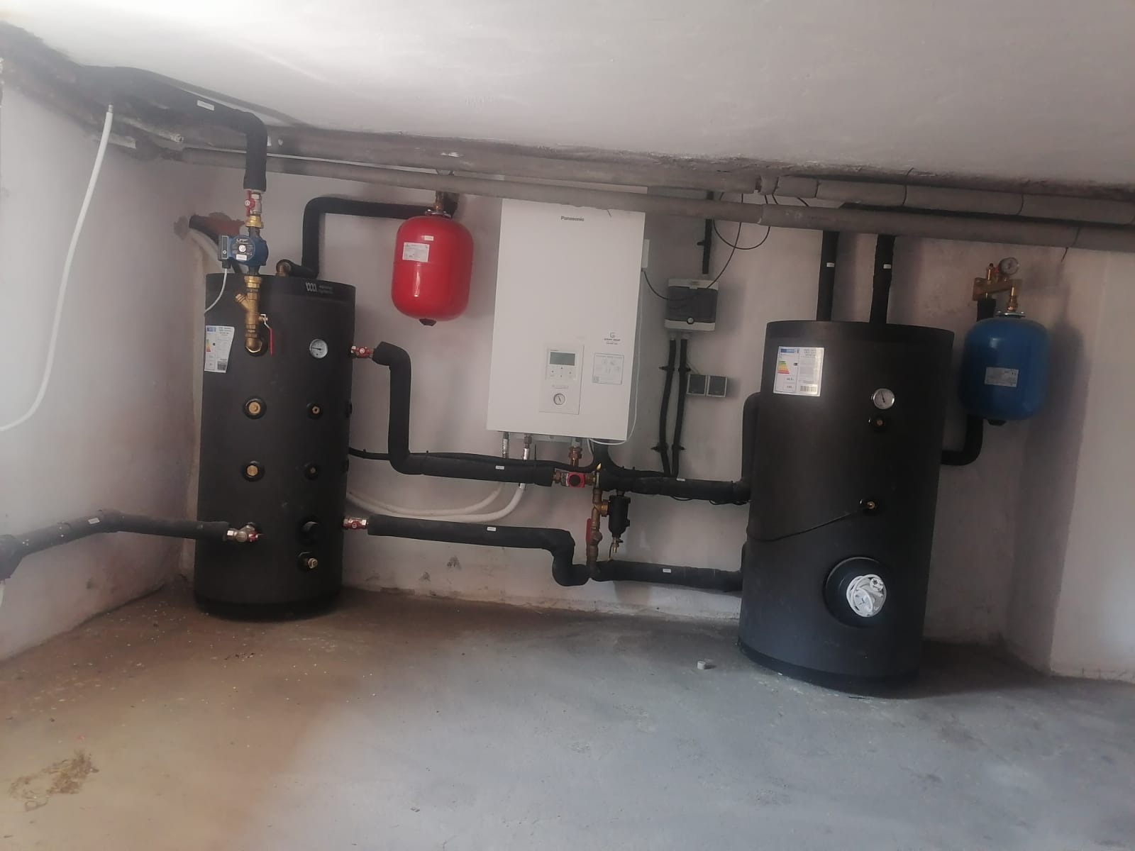 Instalacja w kotłowni: dwa czarne zbiorniki akumulacyjne, biały piec gazowy Panasonic, czerwony i niebieski zbiornik wyrównawczy, rury izolowane i nieizolowane, manometry, zawory, betonowa podłoga...