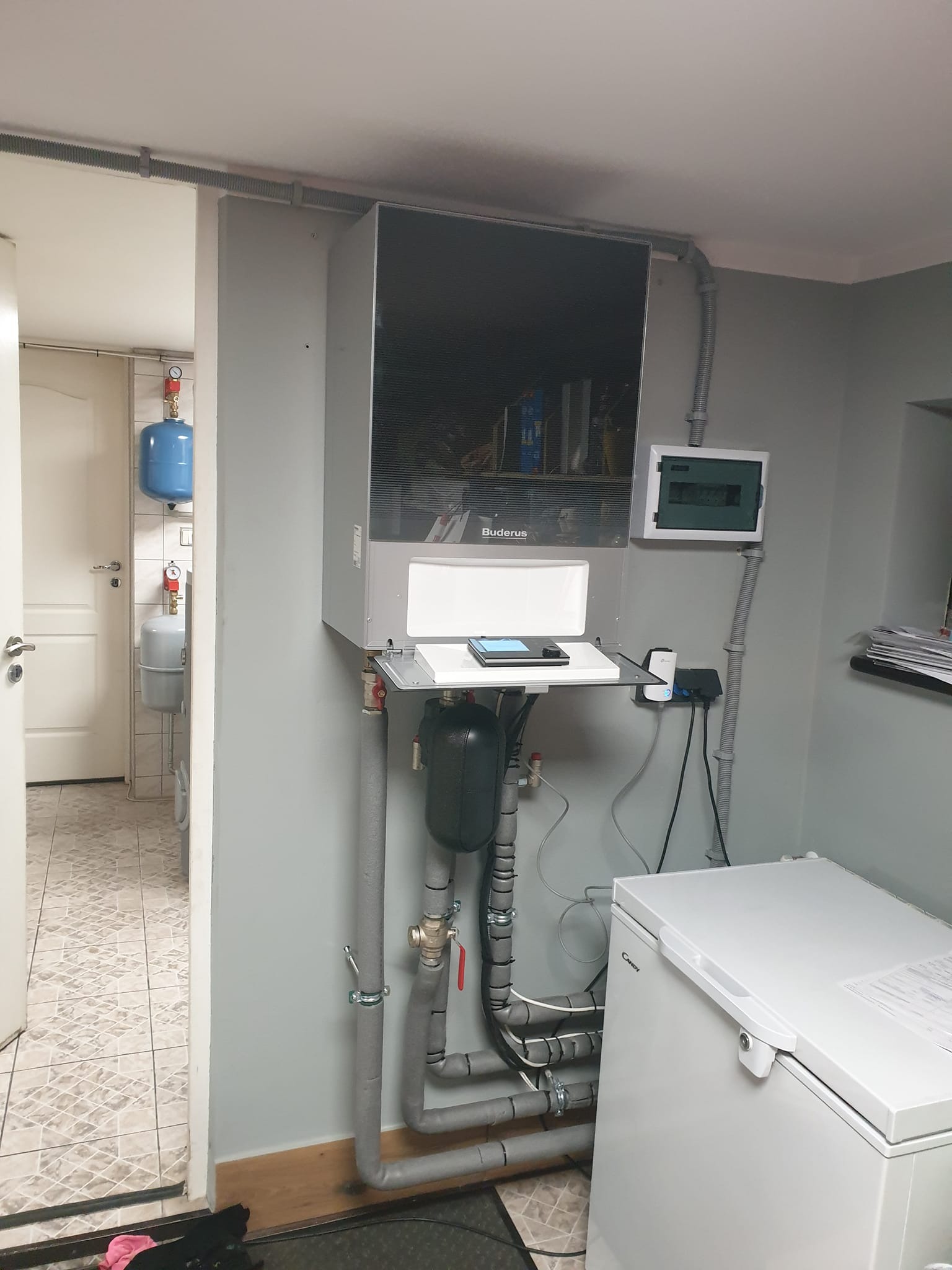 Wnętrze pomieszczenia z zamontowanym urządzeniem Buderus, prawdopodobnie pompą ciepła, z podłączonymi rurami i zbiornikiem wyrównawczym, obok skrzynka elektryczna i zamrażarka skrzyniowa.