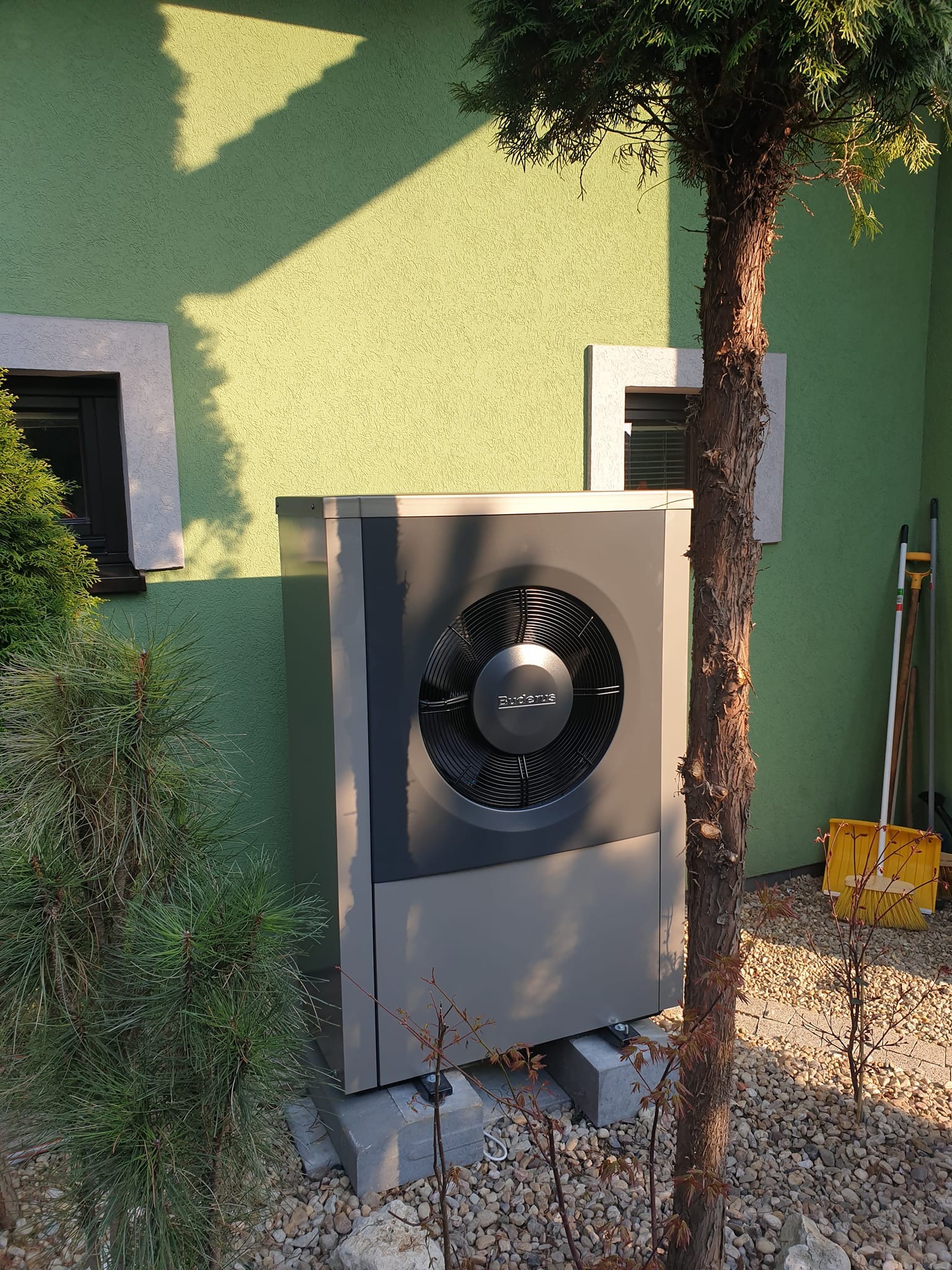 Szara pompa ciepła Buderus na betonowych podstawach, ustawiona przed zieloną ścianą domu z dwoma oknami, częściowo zasłonięta przez iglaste krzewy i małe drzewko.