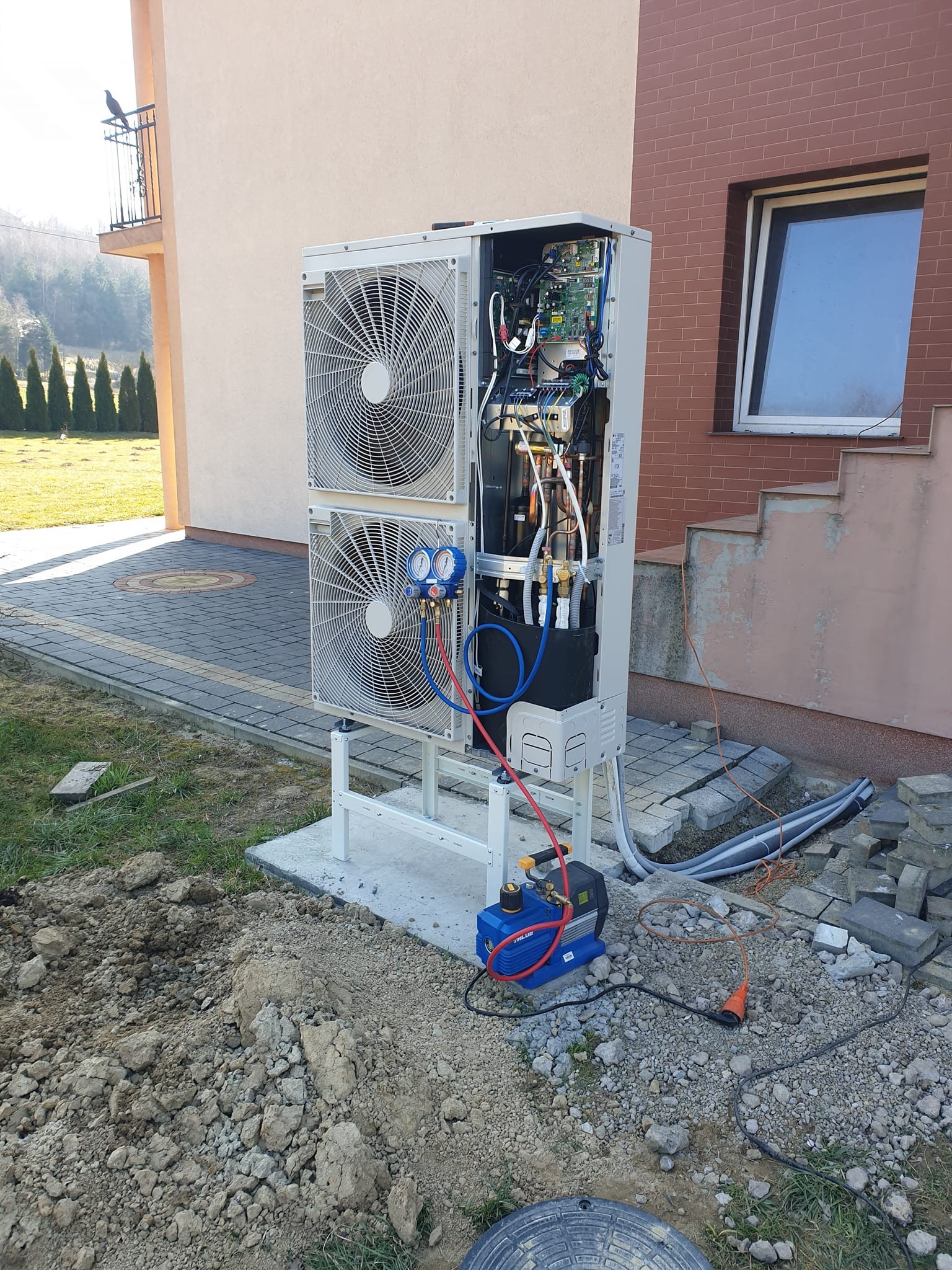 Montaż dwu-wentylatorowej pompy ciepła typu split na zewnątrz budynku, z widocznymi manometrami i pompą próżniową w trakcie instalacji, na tle kostki brukowej i trawnika.