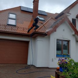 Maclin - Pracownik w kasku czyści dach z brązowej dachówki za pomocą myjki ciśnieniowej. Widoczne panele słoneczne, biała elewacja domu i fragment kostki brukowej.