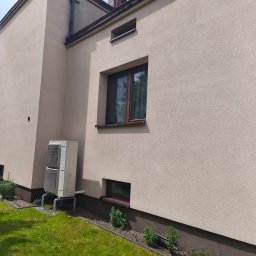 Maclin - Jasna elewacja domu z widocznym klimatyzatorem, brązowymi oknami i rynną. Trawa i rośliny wzdłuż fundamentu.