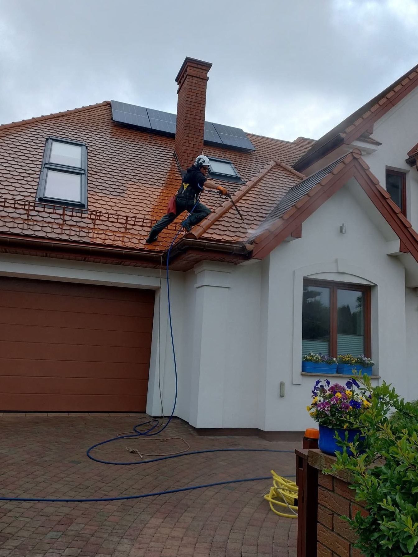 Pracownik w kasku czyści dach z brązowej dachówki za pomocą myjki ciśnieniowej. Widoczne panele słoneczne, biała elewacja domu i fragment kostki brukowej.