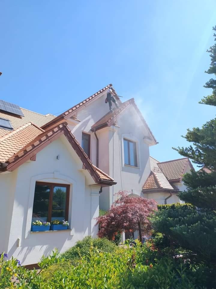 Pracownik w uprzęży, z dachu domu z panelami słonecznymi, spryskuje elewację wodą pod ciśnieniem. Widoczne okna z kwiatami i ozdobne krzewy.