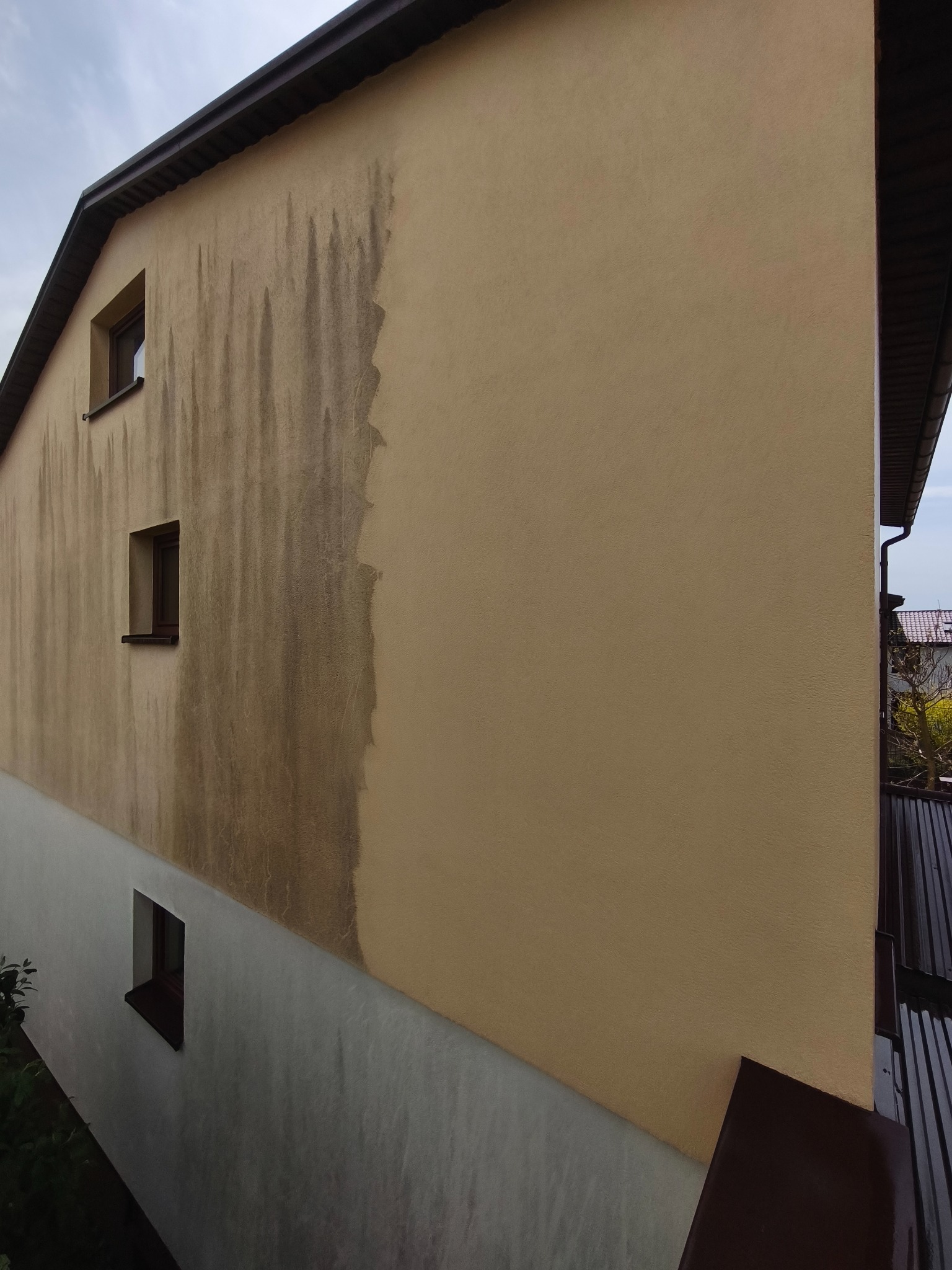 Częściowo umyta elewacja budynku z widocznymi zaciekami i różnicą w kolorze przed i po czyszczeniu. Widoczne trzy okna na bocznej ścianie.