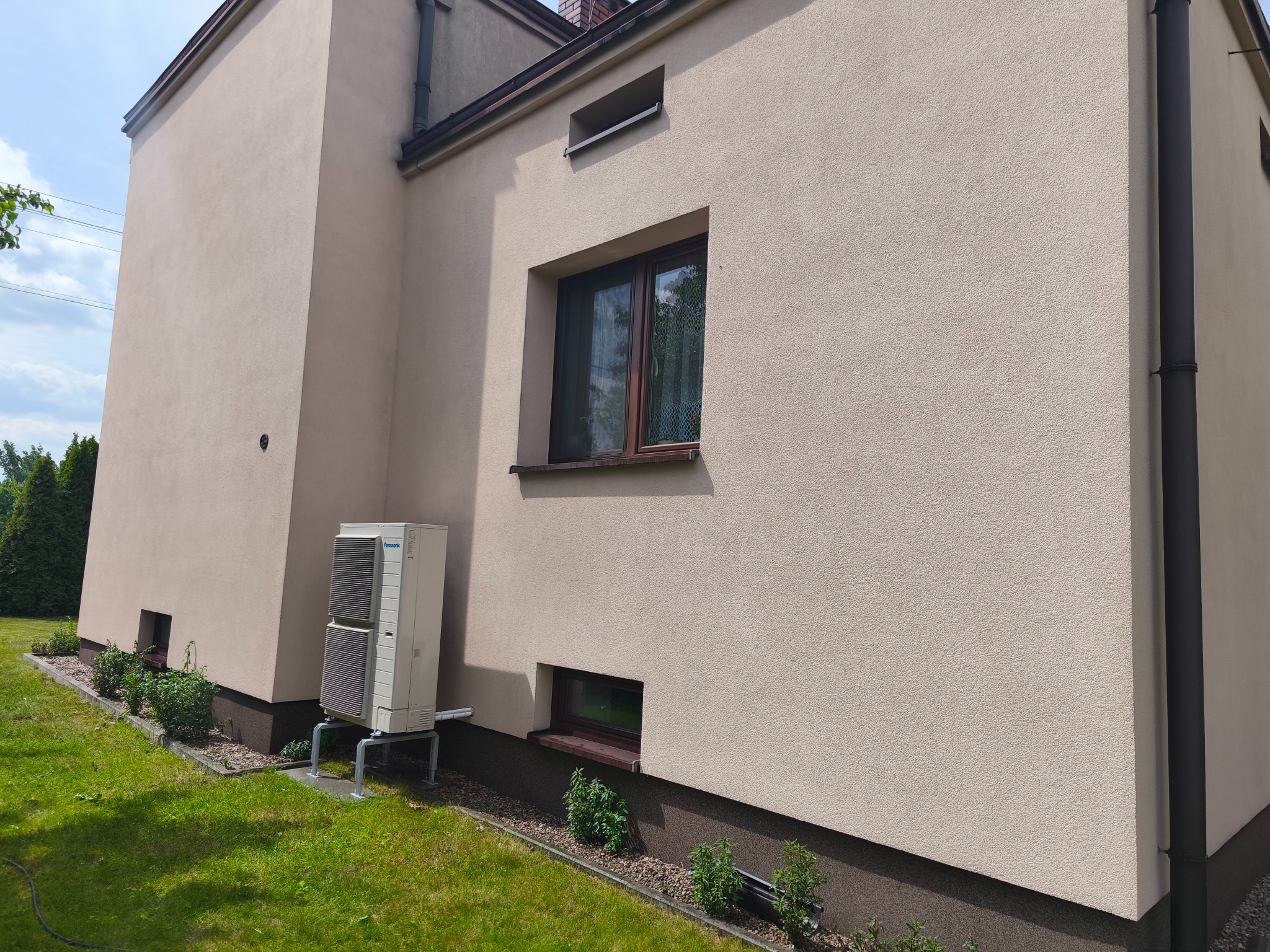 Jasna elewacja domu z widocznym klimatyzatorem, brązowymi oknami i rynną. Trawa i rośliny wzdłuż fundamentu.