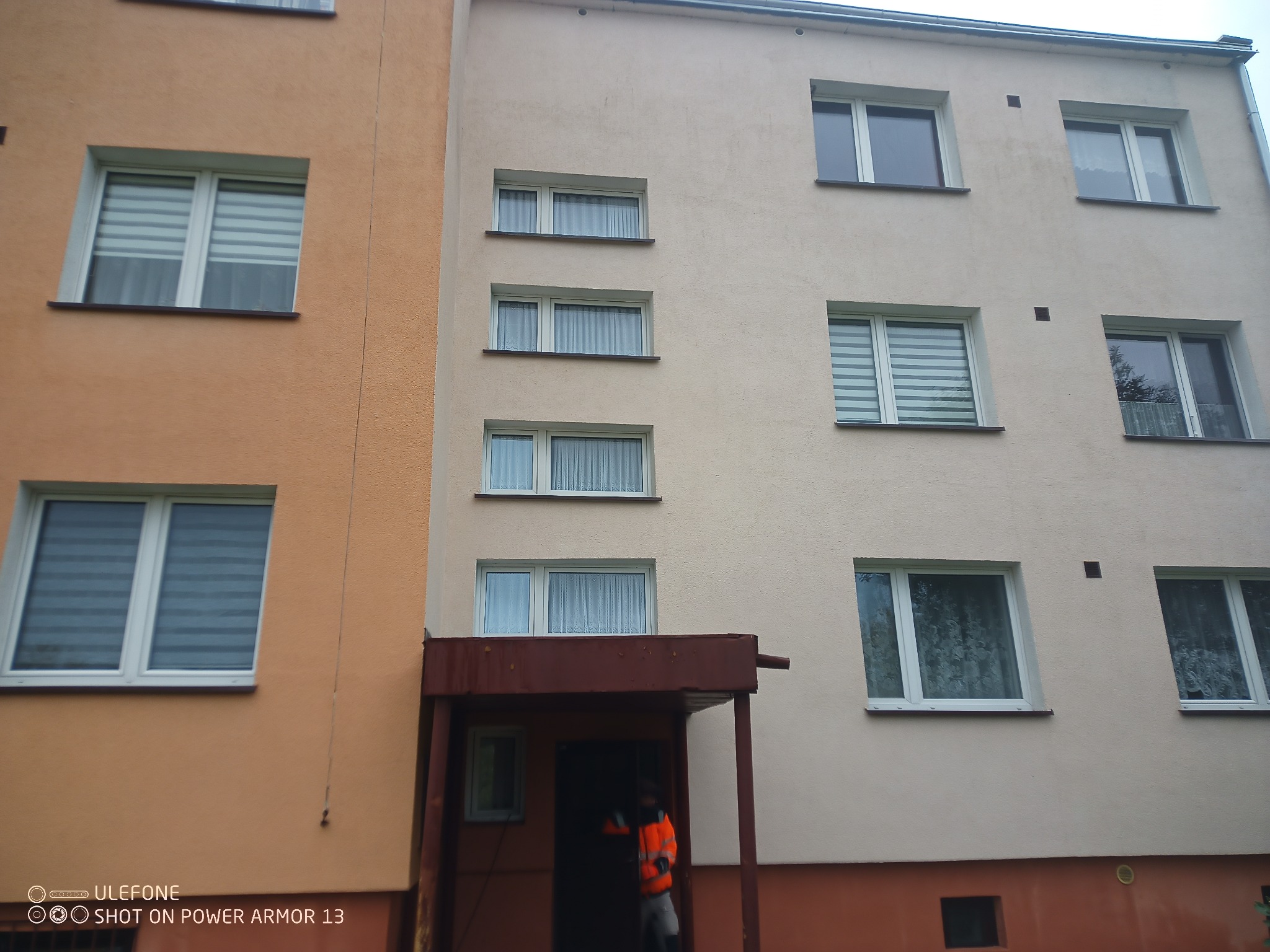 Dwukolorowa elewacja budynku z oknami i zadaszonym wejściem, gdzie widać osobę w pomarańczowej kurtce. Budynek otynkowany, z widocznymi śladami zabrudzeń.