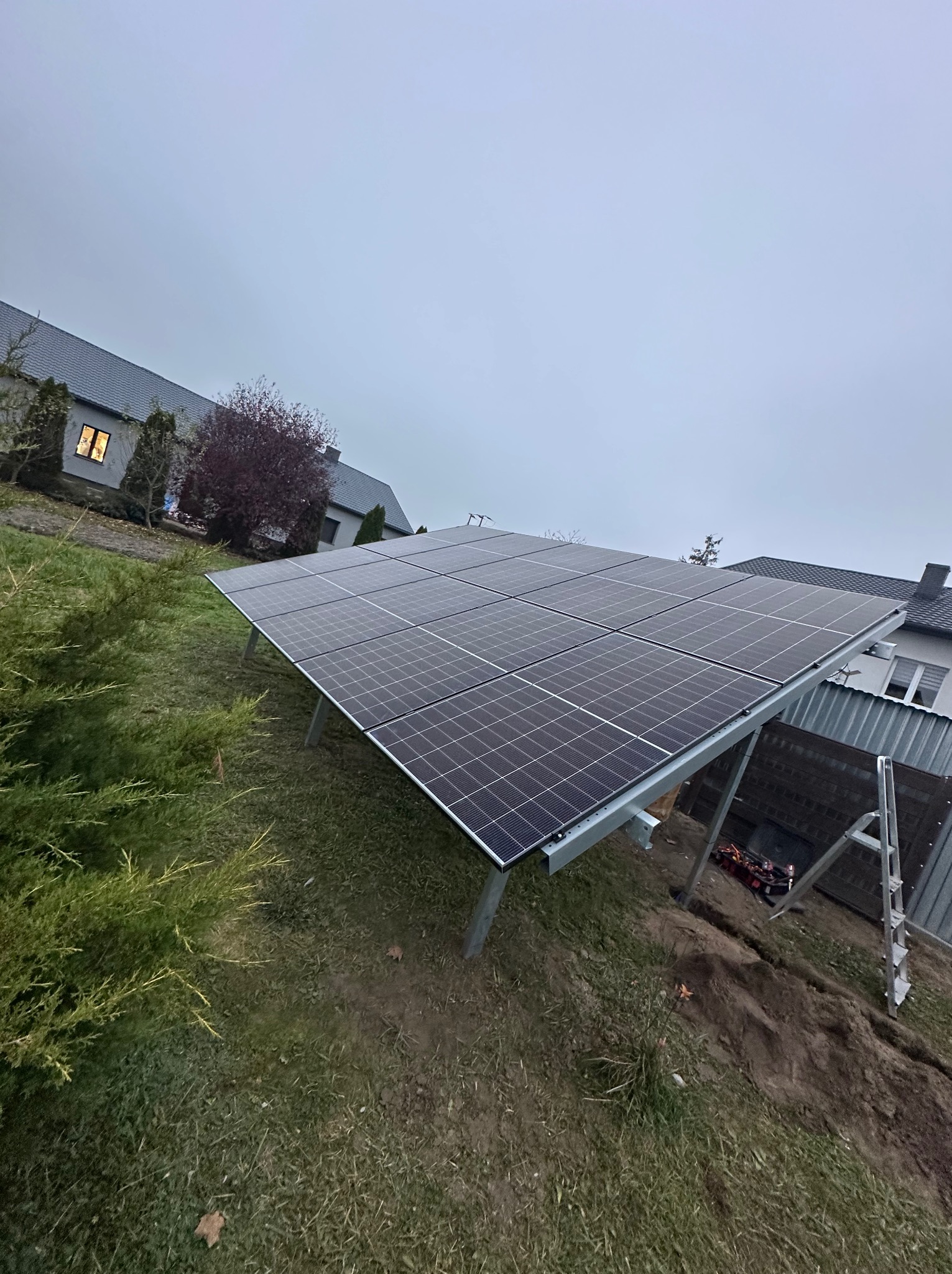 Panel fotowoltaiczny na metalowej konstrukcji, zamontowany na trawniku przy domu jednorodzinnym, widoczna drabina i wykop pod kable.