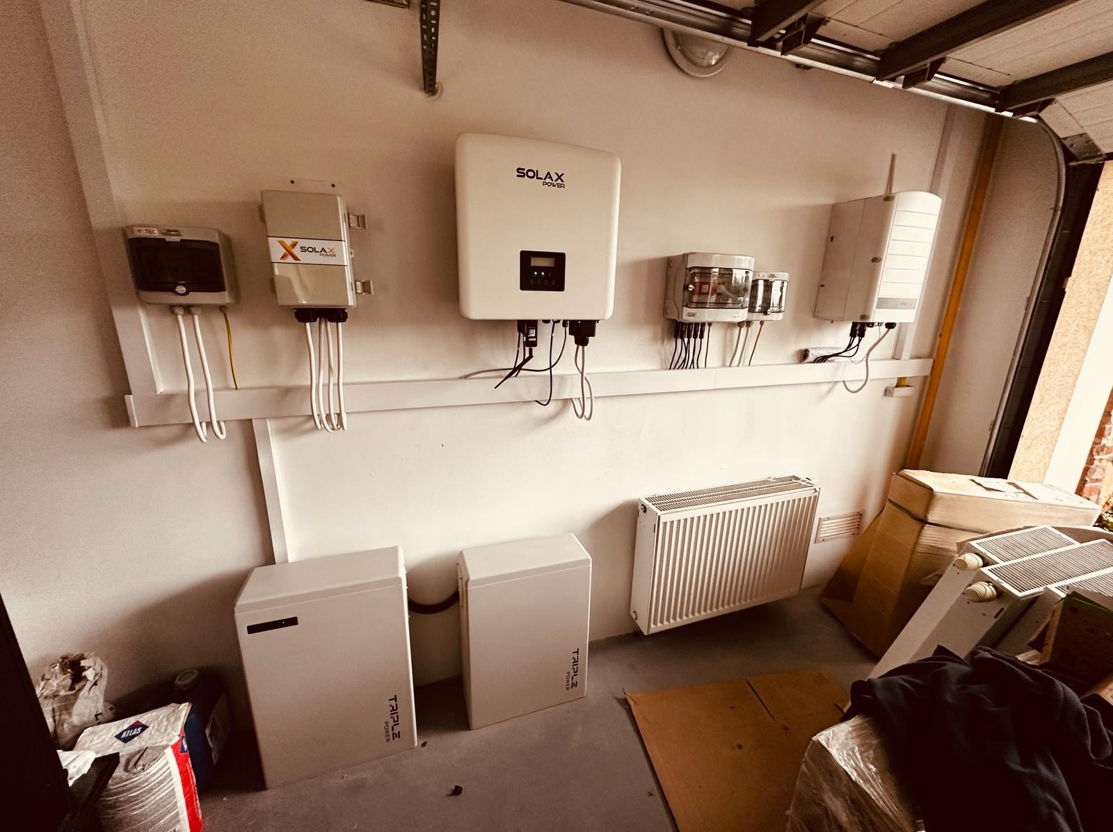 Instalacja fotowoltaiczna w garażu: dwa białe magazyny energii Triple Power, falowniki Solax Power i rozdzielnice zamontowane na ścianie, widoczny grzejnik i fragment bramy garażowej.