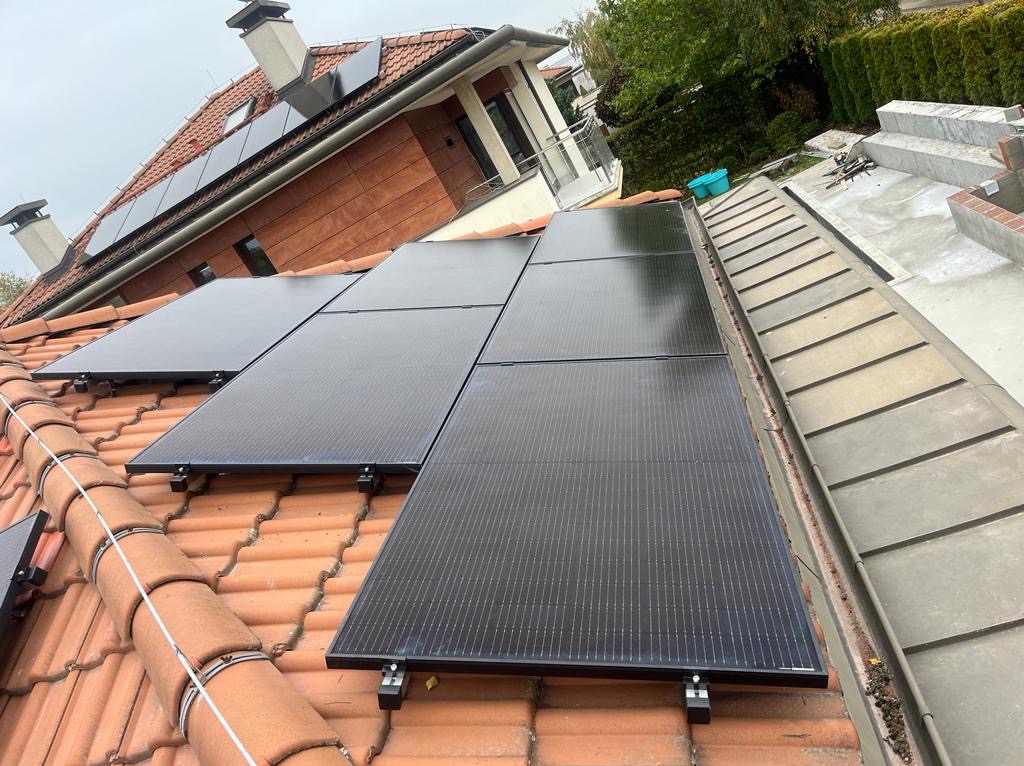 Panele fotowoltaiczne zamontowane na dachu z czerwonej dachówki, widoczne mocowania paneli, w tle budynek mieszkalny z jasną elewacją i fragmentem ogrodu.
