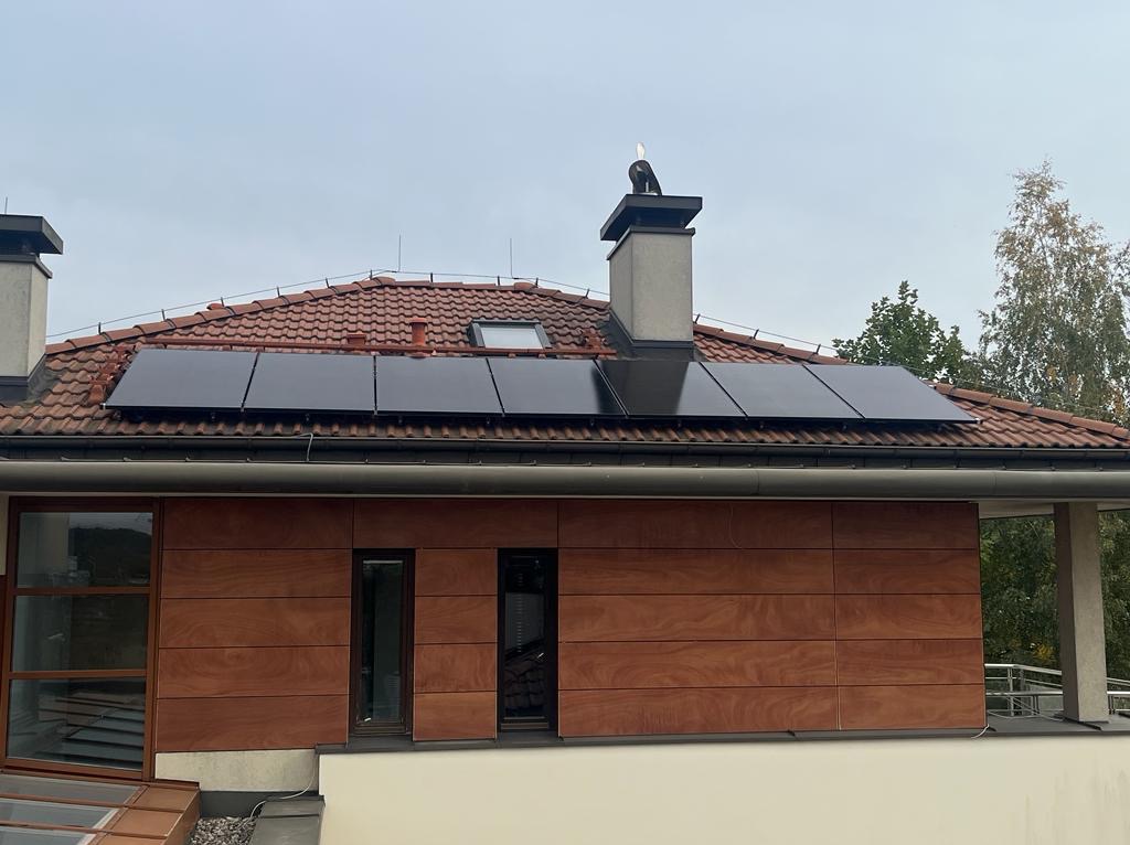 Panele słoneczne zamontowane na dachu domu z czerwoną dachówką, widoczne okno dachowe i komin z ozdobną figurką, elewacja budynku wykończona drewnopodobnymi panelami.