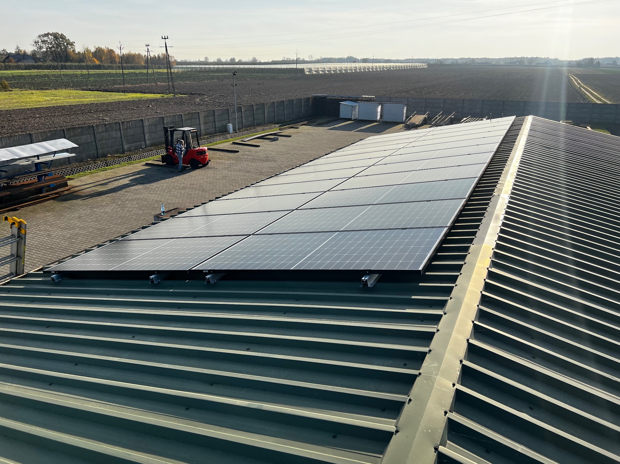 Panele słoneczne zamontowane na dachu blaszanym, widok z góry na instalację z tłem pól uprawnych i wózkiem widłowym w oddali.