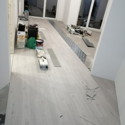 Usługi Elektroniczne Kamil Jabłoński - Wnętrze w trakcie prac: jasne panele podłogowe w trakcie układania, widoczne przycięte fragmenty, narzędzia, puszki z farbą i worki w tle, duże okna balkonowe.