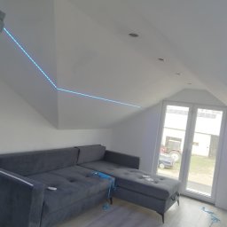Usługi Elektroniczne Kamil Jabłoński - Instalacja oświetlenia LED na poddaszu, widoczny fragment pracownika, szara sofa, okno z widokiem na traktor i budynek gospodarczy.