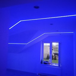 Usługi Elektroniczne Kamil Jabłoński - Niebieskie oświetlenie LED zamontowane wzdłuż krawędzi sufitu i ścian w pokoju z oknem.