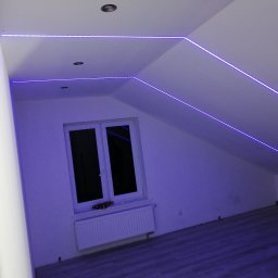 Usługi Elektroniczne Kamil Jabłoński - Pokój na poddaszu z oknem i grzejnikiem, oświetlony niebieskimi paskami LED zamontowanymi wzdłuż krawędzi ścian i sufitu, tworząc geometryczne wzory.