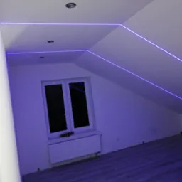 Pokój na poddaszu z oknem i grzejnikiem, oświetlony niebieskimi paskami LED zamontowanymi wzdłuż krawędzi ścian i sufitu, tworząc geometryczne wzory.