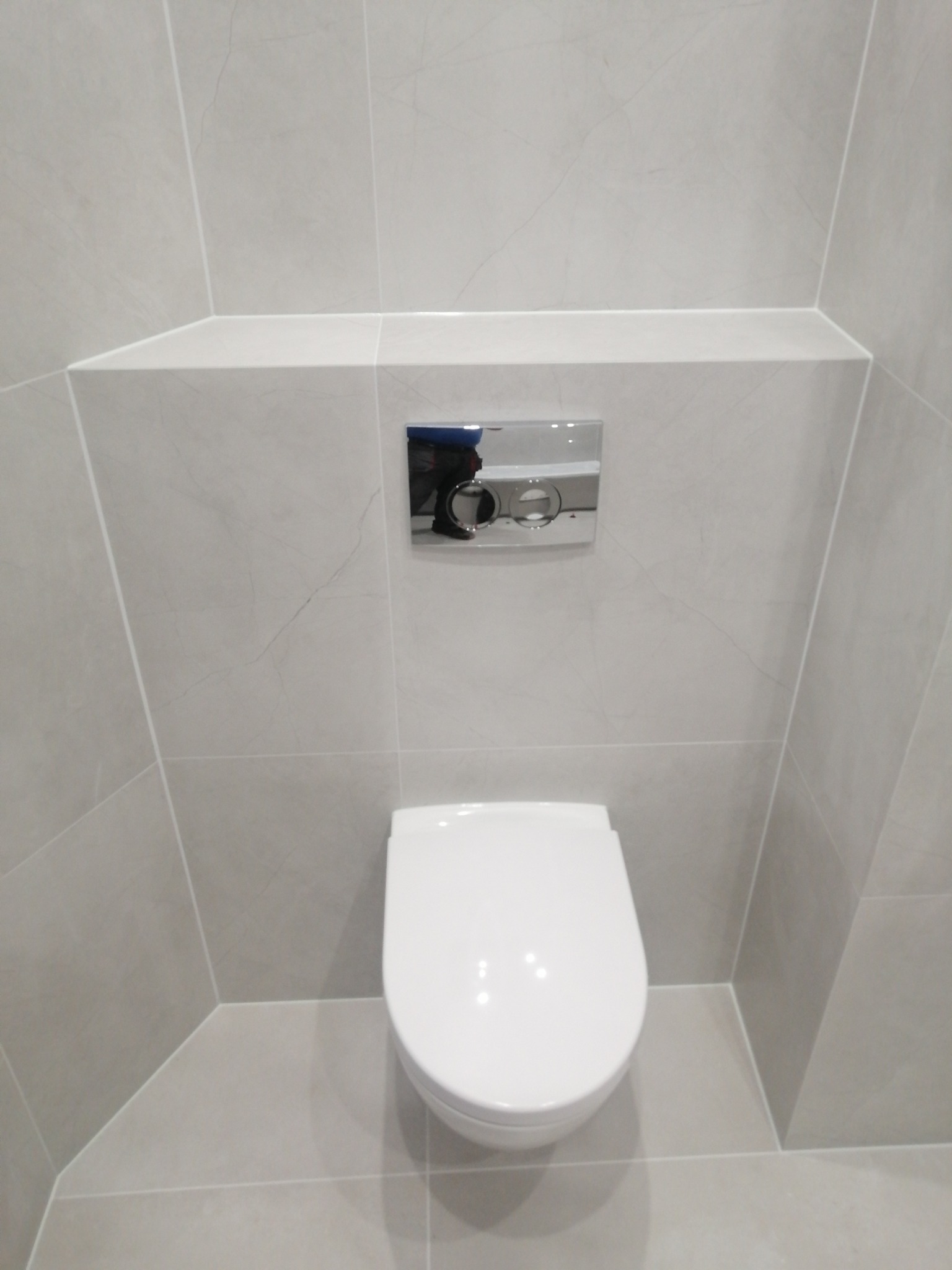 Podwieszana toaleta w nowoczesnej łazience wykończonej szarymi płytkami imitującymi marmur, z chromowanym przyciskiem spłukującym.