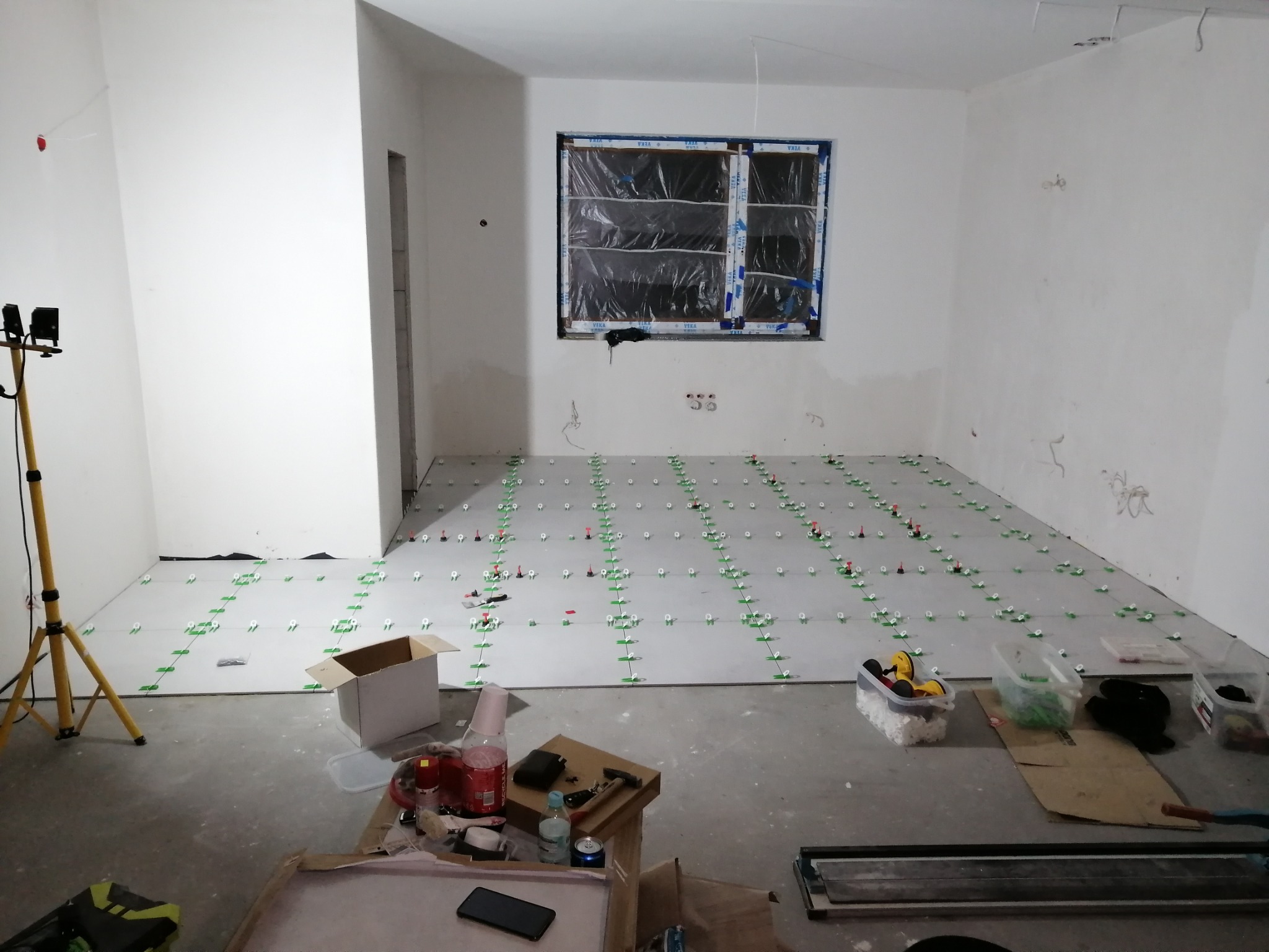 Układanie szarych płytek podłogowych z użyciem zielonych krzyżyków dystansowych w pomieszczeniu z oknem zabezpieczonym folią, widoczne narzędzia i lampa na statywie.