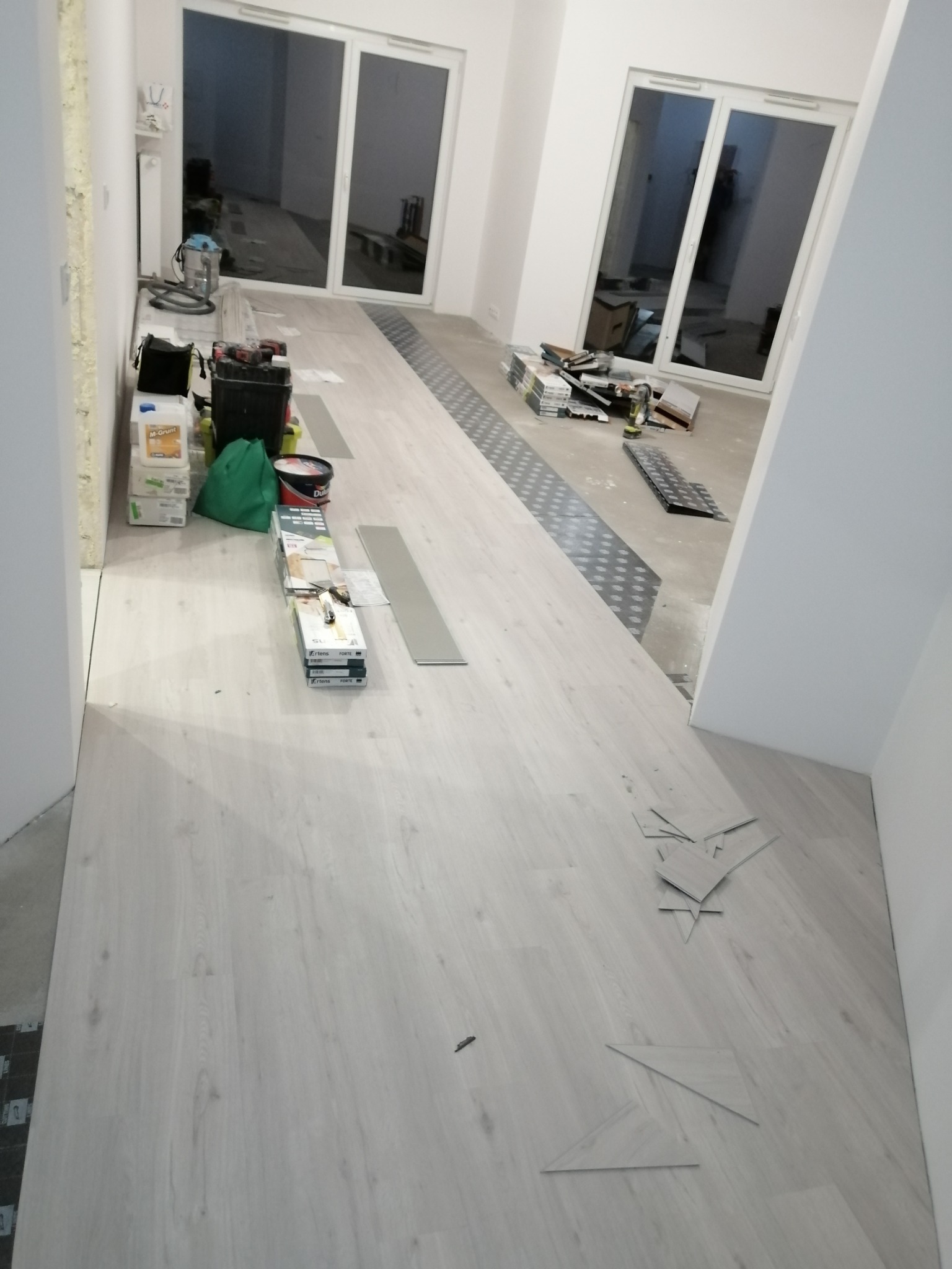 Wnętrze w trakcie prac: jasne panele podłogowe w trakcie układania, widoczne przycięte fragmenty, narzędzia, puszki z farbą i worki w tle, duże okna balkonowe.