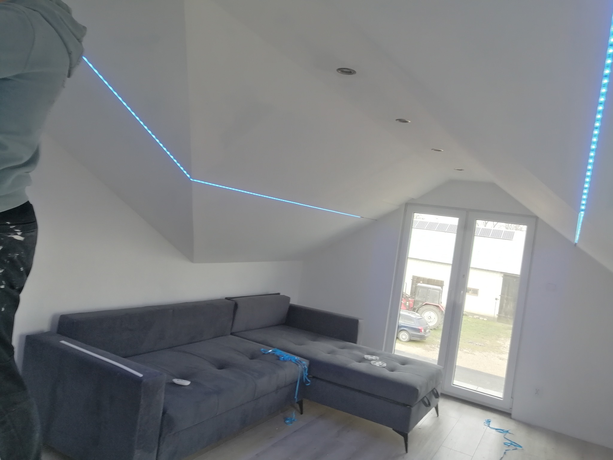 Instalacja oświetlenia LED na poddaszu, widoczny fragment pracownika, szara sofa, okno z widokiem na traktor i budynek gospodarczy.