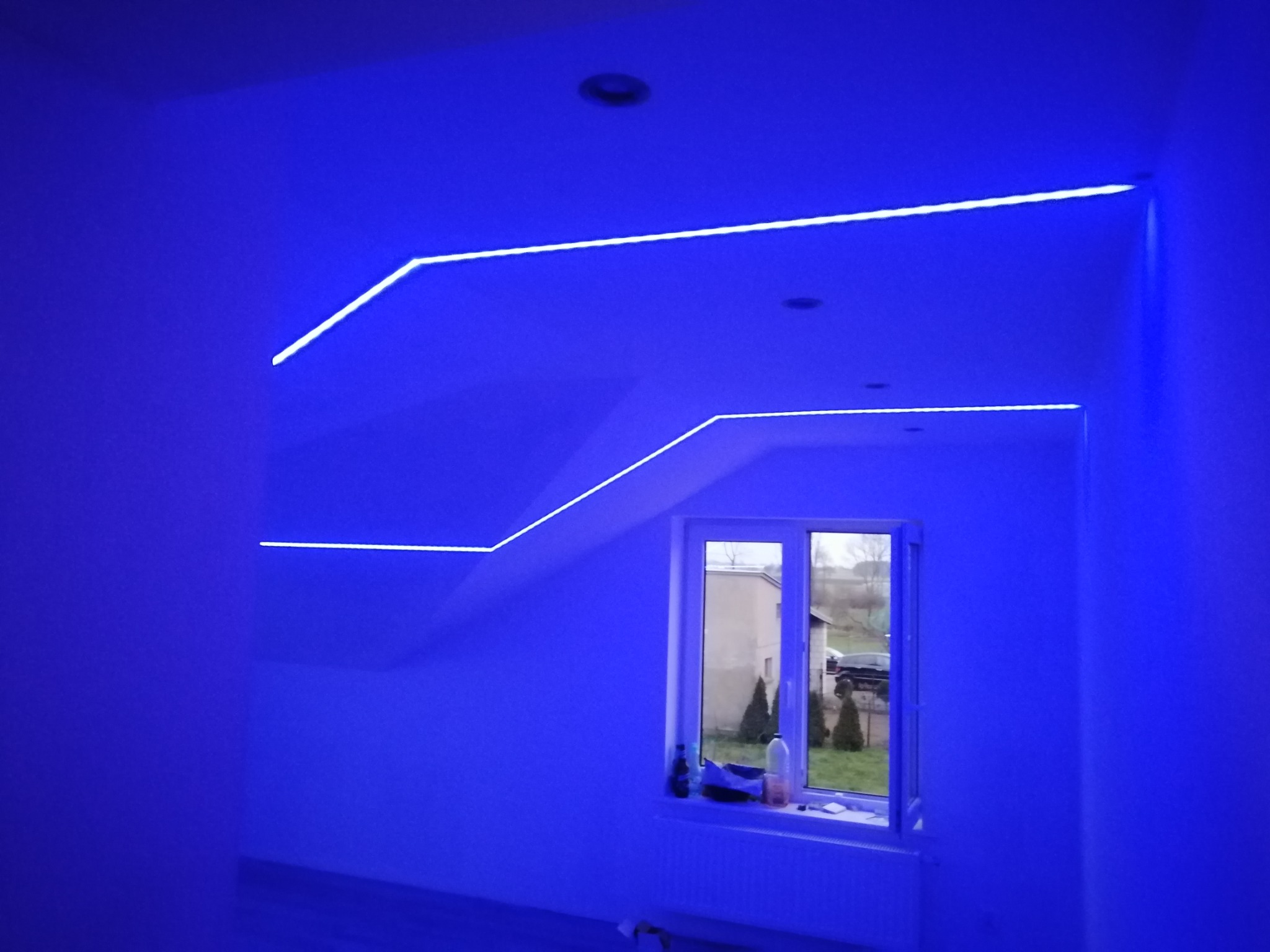 Niebieskie oświetlenie LED zamontowane wzdłuż krawędzi sufitu i ścian w pokoju z oknem.