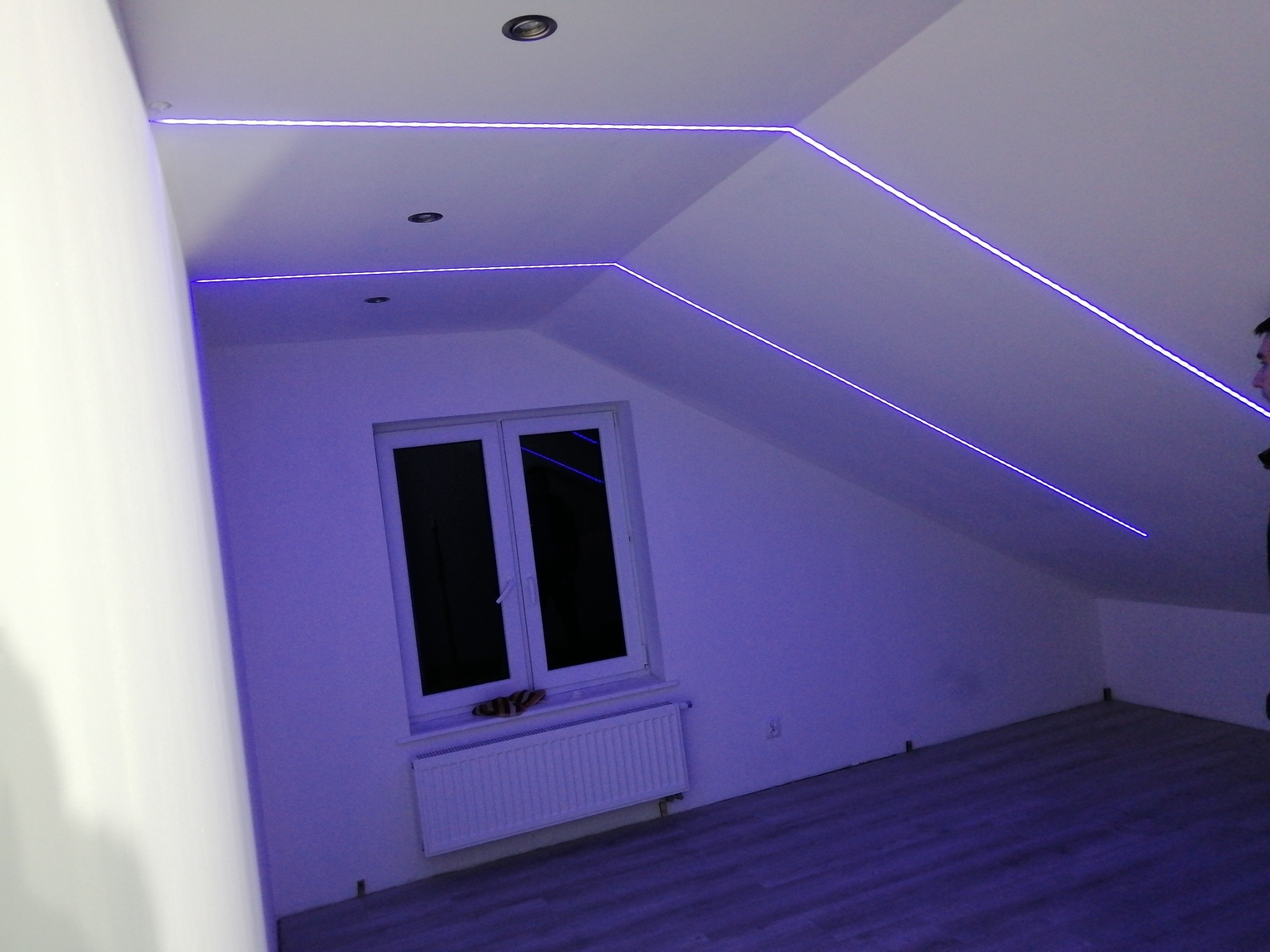 Pokój na poddaszu z oknem i grzejnikiem, oświetlony niebieskimi paskami LED zamontowanymi wzdłuż krawędzi ścian i sufitu, tworząc geometryczne wzory.