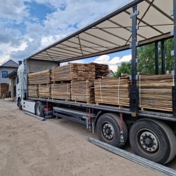 WOOD-FERT - Ciężarówka DAF z naczepą załadowana paletami z pociętym drewnem, prawdopodobnie deski, na tle budynku mieszkalnego i pochmurnego nieba.