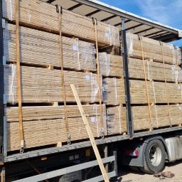 WOOD-FERT - Ciężarówka z czerwonym ciągnikiem siodłowym załadowana paletami z drewnem kominkowym, zabezpieczonym folią, na tle błękitnego nieba i budynku magazynowego.