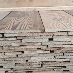WOOD-FERT - Stos równo ułożonych, prostokątnych desek z widocznym usłojeniem, o różnych odcieniach brązu i szarości, przygotowanych do transportu lub składowania.