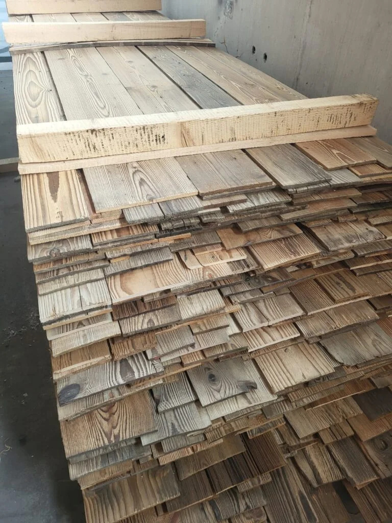 Stos drewnianych desek o nieregularnych krawędziach, ułożonych warstwami i zabezpieczonych belkami, ukazujący naturalny rysunek drewna i jego zróżnicowaną kolorystykę.