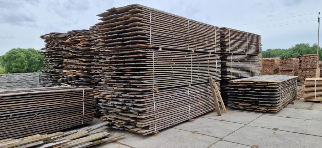 Stosy desek ułożone na paletach i zabezpieczone sznurkiem, widoczne na placu składowym z betonową nawierzchnią i zielenią w tle.