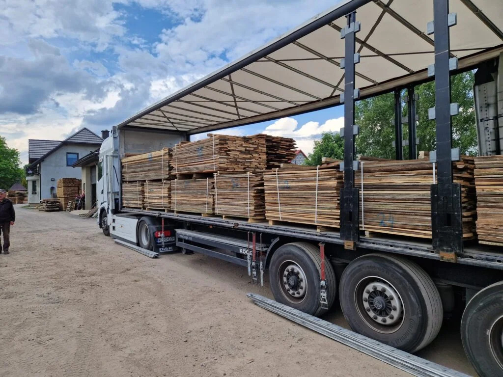 Ciężarówka DAF z naczepą załadowana paletami z pociętym drewnem, prawdopodobnie deski, na tle budynku mieszkalnego i pochmurnego nieba.